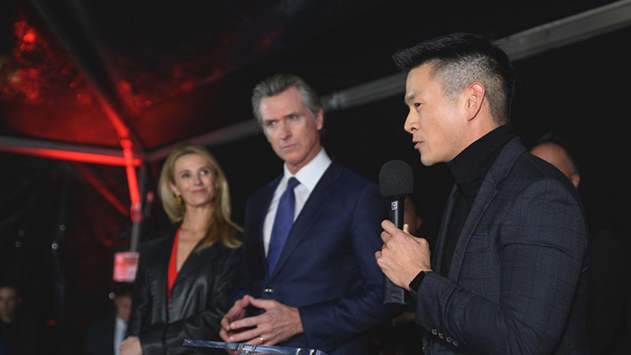 Assemblymember Evan Low holding a microphone standing at a podium next to Governor Newsom and Jennifer Siebel Newsom